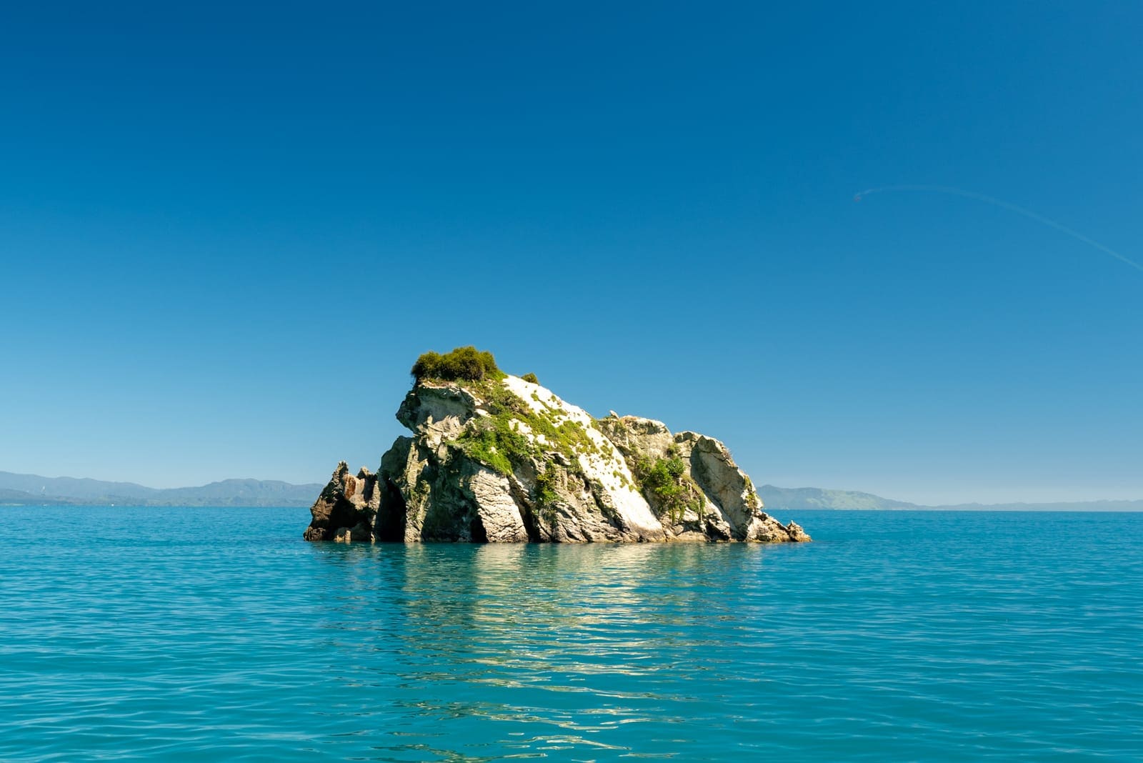 A single rugged rock islet topped with green scrub rising from calm turquoise water in Tasman Bay, distant hills on the horizon.